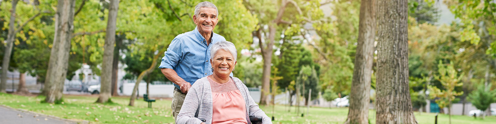 A senior man pushing his wife in a wheelchair outside at a nursing home