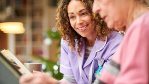 A senior woman wearing a pink shirt looking down at a photo album while sitting beside her female caregiver who is wearing purple scrubs.